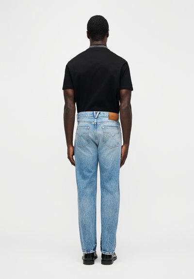 Vyras dėvi šviesiai mėlynas tiesaus silueto džinsus su „Versace“ etikete, juodą trumpomis rankovėmis marškinį ir juodus batus, stovintis nugara į priekį ant vienspalvio fono.