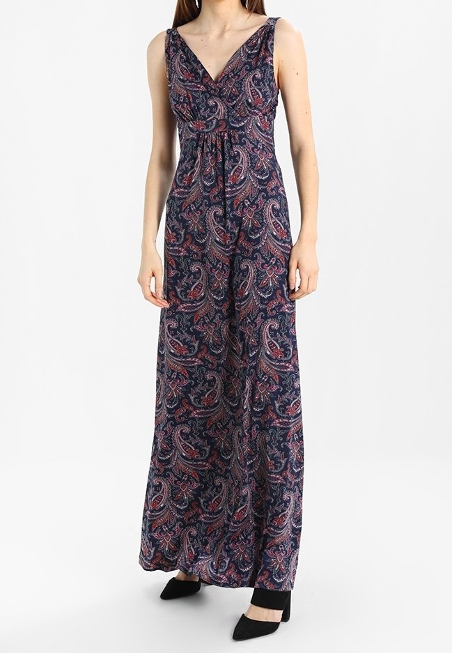 Femme portant une robe maxi sans manches bleu marine avec des motifs cachemire rouges et roses, associée à des chaussures noires à talons.