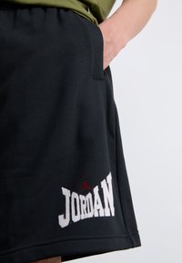 Shorts noirs avec une poche sur le côté, présentant un logo "JORDAN" blanc et un petit emblème Jumpman rouge, tenus par la main d'une personne.