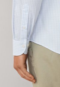 Desna ruka osobe koja nosi svijetloplavu košulju na karirane uzorke s dugmetom na manžetu i bež hlačama, ruka opušteno uz tijelo protiv neutralne pozadine.