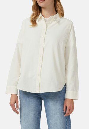 Kobieta ubrana w luźną, białą, długorękawową koszulę z kołnierzykiem i guzikami, w połączeniu z jasnoniebieskimi dżinsami, stojąca na jednolitym tle.