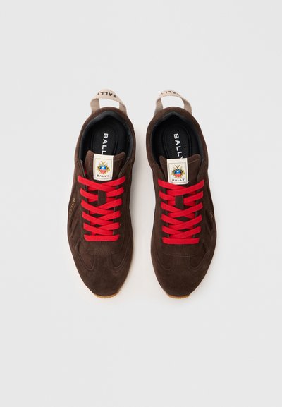 Tênis de camurça castanho com atacadores vermelhos, apresentando um logótipo na língua e uma sola de borracha texturizada. Design simples e aerodinâmico.