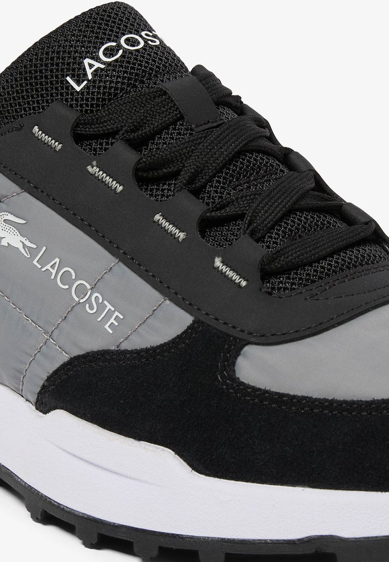 Baskets Lacoste noires et grises avec une languette en maille, un laçage à l'avant, un logo blanc et une semelle texturée visible.