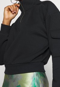 Svart zip-up sweatshirt med hög krage, ribbade ärmslut och sidoficka. Mjukt tyg med en avslappnad passform. Matcher med en grön mönstrad kjol.