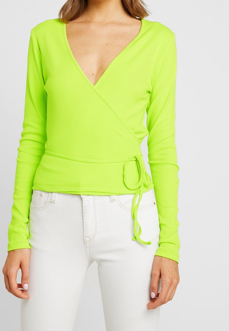 Neongrön wrap-top med långa ärmar, djup V-ringning, ribbad textur och justerbar snörning i midjan, tillsammans med vita jeans.
