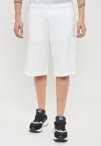 Shorts bianchi in cotone con vita elastica, dotati di tasche laterali e un dettaglio con logo testurizzato sulla gamba sinistra, abbinati a sneakers nere.