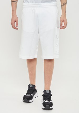 Hvide bomuldsshorts med elastisk talje, der har sidelommer og en tekstureret logo-detalje på venstre ben, parret med sorte sneakers.