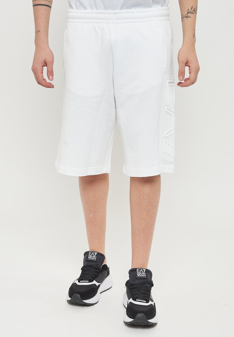 Shorts bianchi in cotone con vita elastica, dotati di tasche laterali e un dettaglio con logo testurizzato sulla gamba sinistra, abbinati a sneakers nere.