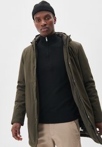 Olivgrüne Parka mit einem Reißverschluss vorne, verstellbarem Kapuzen und strukturiertem schwarzen Rollkragenpullover darunter; getragen mit beigen Hosen.