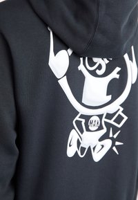 Svart huvtröja med en vit grafisk design som har en tecknad karaktär och texten "MAX". Materialet verkar mjukt med en avslappnad passform och snörning i huvan.