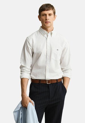 Homme portant une chemise blanche à rayures avec manches retroussées, pantalon sombre à fines rayures, ceinture marron, tenant une veste bleu clair dans la main gauche.