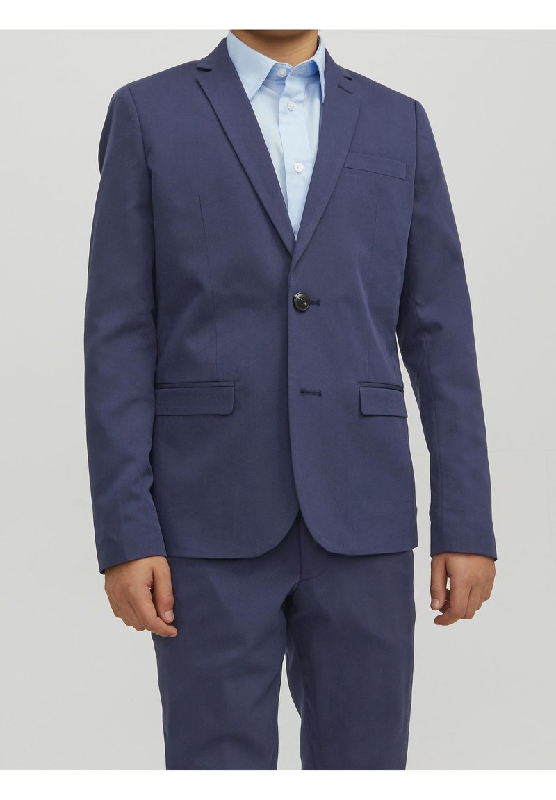 Granatowy garnitur z pojedynczym zapięciem na guziki, dwoma kieszeniami z przodu i klapami z wycięciem. Noszony na jasnoniebieskiej koszuli z kołnierzykiem. Gładka faktura.