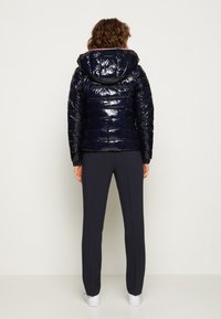 Kobieta stoi przodem do ściany, ubrana w błyszczący granatowy puchowy kurtkę z kapturem, ciemne proste spodnie i białe sneakersy.