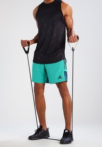 Svart ärmlös träningsskjorta, teal shorts med mörka accenter, håller i motståndsband, i kombination med svarta träningsskor.