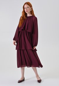 Abito in chiffon bordeaux con maniche lunghe, design a strati, vita elasticizzata e texture morbida e fluida, abbinato a mocassini scuri.