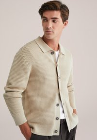 Cardigan texturat bej cu guler, având un închidere cu nasturi, manșete cu nervuri și o croială relaxată, purtat peste o cămașă albă.