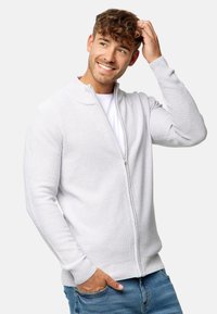 Cardigan de cremallera gris claro con un tejido texturizado, puños y dobladillo acanalados, usado sobre una camisa blanca, acompañado de jeans azules.