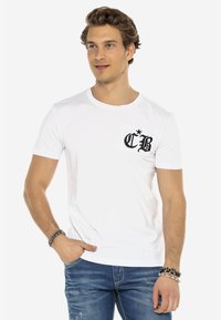 Weißes Baumwoll-T-Shirt mit einer Brusttasche und einem schwarzen grafischen Design. Kurze Ärmel und runder Ausschnitt. Kombination mit blauen Jeans.
