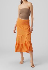 Žena oblečená v hnědém topu bez ramínek, midi sukni s oranžovým vzorem a volánky, a béžových sandálech na podpatku, stojící na světle šedém pozadí.