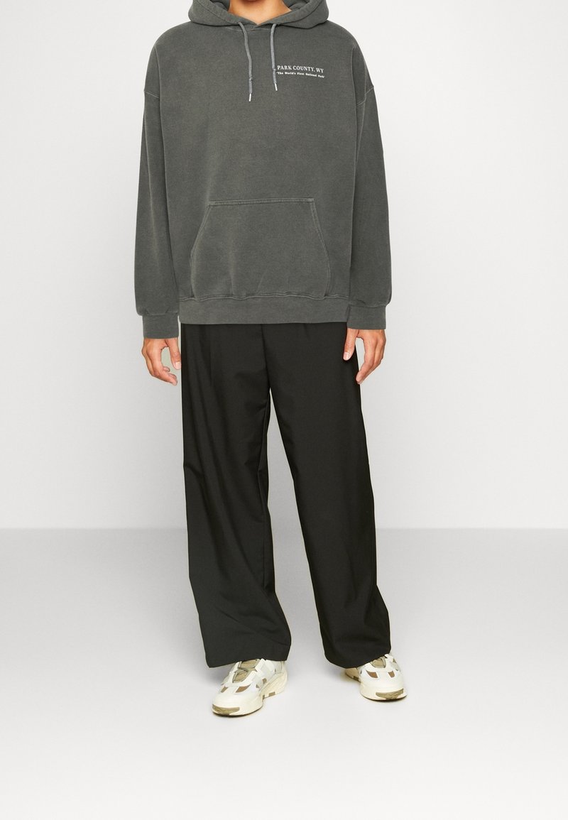 Felpa con cappuccio grigio carbone, dotata di tasca a marsupio e cordino, abbinata a pantaloni a gamba larga neri e sneakers chiare.