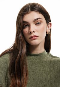 Olijfgroene, gestructureerde top met een hoge hals. Model draagt een kleine gouden ringoorbel. Het haar is lang en golvend, vormt een omlijsting rond het gezicht.