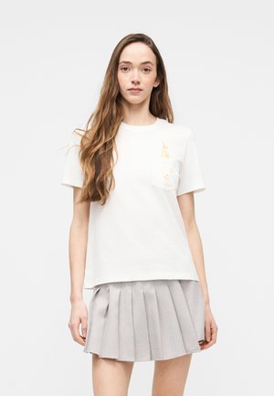 Giovane donna con lunghi capelli castani che indossa una maglietta bianca a maniche corte con piccoli motivi gialli e una gonna grigia a pieghe.