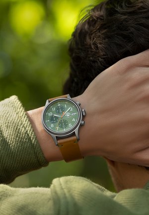 Runde Uhr mit grünem Zifferblatt, weißen Ziffern, silbergrauem Gehäuse und braunem Lederarmband. Verfügt über mehrere Hilfszifferblätter und eine Datumsfunktion.