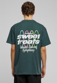 Mørkegrøn bomulds t-shirt med hvid og farverig tekst på ryggen, med sætningen "søde lækkerier" og et dekorativt design.