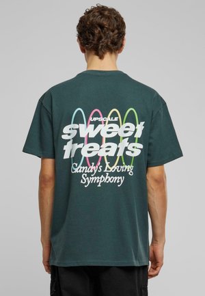 Mørkegrøn bomulds t-shirt med hvid og farverig tekst på ryggen, med sætningen "søde lækkerier" og et dekorativt design.