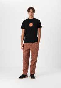 Black cotton t-shirt features a colorful circular graphic. Brown patterned trousers with floral designs. Black sneakers complete the outfit.