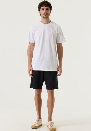 Man staat, draagt een effen wit T-shirt met korte mouwen, zwarte knielange korte broek en beige sneakers tegen een lichte achtergrond.