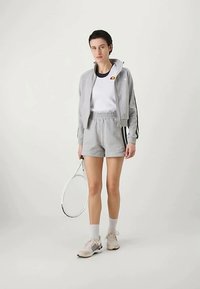Γυναίκα σε γκρι αθλητική ενδυμασία holding a tennis racket, φ wearing a cropped jacket, σορτσάκια, λευκό πουκάμισο, κάλτσες και μπεζ αθλητικά παπούτσια, standing against a plain background.