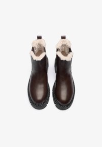 Bottines en cuir marron avec une doublure légère et pelucheuse, bout arrondi et semelle texturée noire. Conçues avec des languettes pour un enfilage facile.
