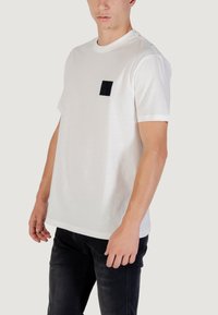Camiseta de algodón blanca con mangas cortas y un bolsillo rectangular negro en el lado izquierdo del pecho. Corte recto en el dobladillo y textura suave.