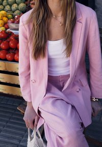 Nainen istuu penkillä, yllään vaaleanpunainen puku ja valkoinen toppi, pitäen käsissään kudottua laukkua, taustalla tuoreita tomaatteja ja muita tuotteita.