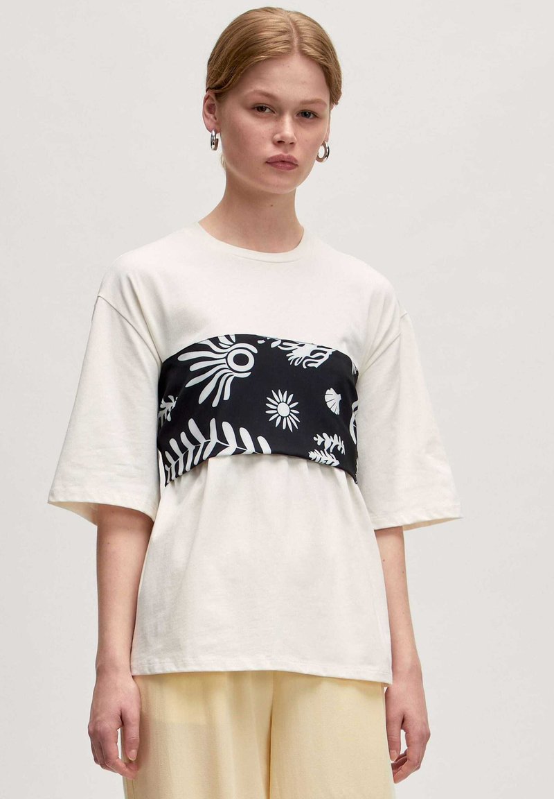 Witte t-shirt met korte mouwen en een zwarte patroonband over de borst met witte bladeren en abstracte ontwerpen.