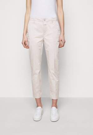 Osoba nosząca jasnobeżowe krótkie spodnie i białe trampki, stojąca na jednolitym białym tle.