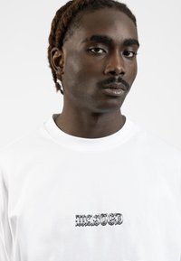 Camiseta de algodón blanca con un gráfico negro de "WASTED" en una fuente gótica en negrita en el pecho. Diseño de cuello redondo simple.