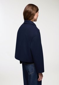 Giacca corta blu navy con colletto a risvolto e maniche larghe, realizzata in tessuto testurizzato, abbinata a jeans in denim scuri.