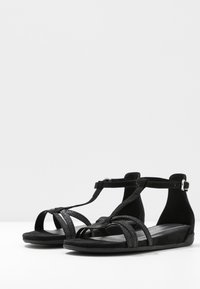Schwarze Wildledersandalen mit T-Riemen-Design, strukturierten Akzenten und offener Zehe. Flache Sohle sorgt für Komfort mit verstellbarem Knöchelriemen.