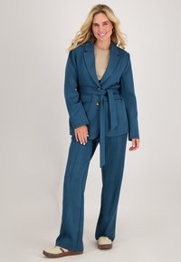 Teal blazer met een gestrikt taille, gecombineerd met bijpassende teal broek. Zachte stof met een lichte glans. Laag uitgesneden sneakers met roomkleurige en bordeauxrode accenten.