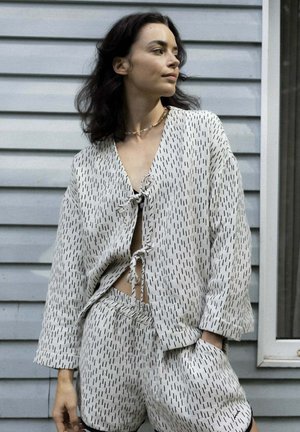 Woman with dark hair wears a light matching outfit with black dashes, consisting of a loose tie-front top and shorts, standing by a gray siding wall.