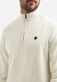 Lichtbeige ritsvest van zacht materiaal; heeft een hoge kraag, subtiele textuur en een klein hexagonaal logo op de borst.