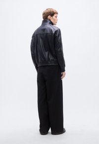 Fekete bőrdzseki magas gallérral, bordázott mandzsettákkal és szegéllyel. Laza fekete nadrág fölött viselve, hátulnézetből.