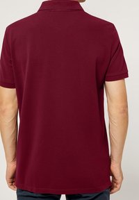 Burgundská polo košile, krátké rukávy, materiál z bavlněné směsi, klasický límec, hladká textura a rovný lem s bočními štěrbínami.