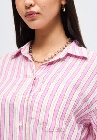 Γυναίκα που φοράει ροζ και λευκό ριγέ πουκάμισο με κουμπιά και τσέπη μπροστά, ασημένιο κολιέ με χάντρες σε σχήμα μπάλας και ασημένια κρίκους σκουλαρίκια.