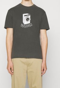 T-shirt grigio scuro con scollatura rotonda, caratterizzata da una grafica bianca di una lavatrice e la scritta "VOLCOM" in carattere stilizzato. Maniche corte.