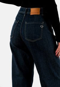 Vaqueros de mezclilla azul oscuro con cintura alta, que cuentan con una etiqueta de cuero marrón grabada con "PLEASE" y un acento en forma de corazón plateado en el bolsillo.