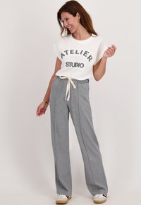 Vrouw die glimlacht, staat, draagt een witte "ATELIER STUDIO" t-shirt, grijze joggingsbroek met koordje en witte sneakers op een effen lichte achtergrond.