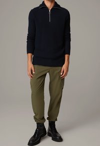 Pull en maille bleu marine avec un col zippé, associé à un pantalon cargo vert olive et des bottines noires à lacets. Tissu texturé avec des détails côtelés.